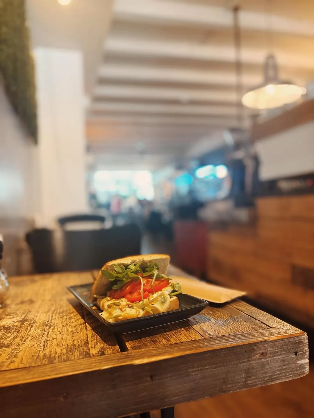 A close-up shot of a sandwich filled with greens, sliced tomatoes, and a creamy ingredient, served on a rectangular black plate on a wooden table. The background shows a blurred cafe interior.