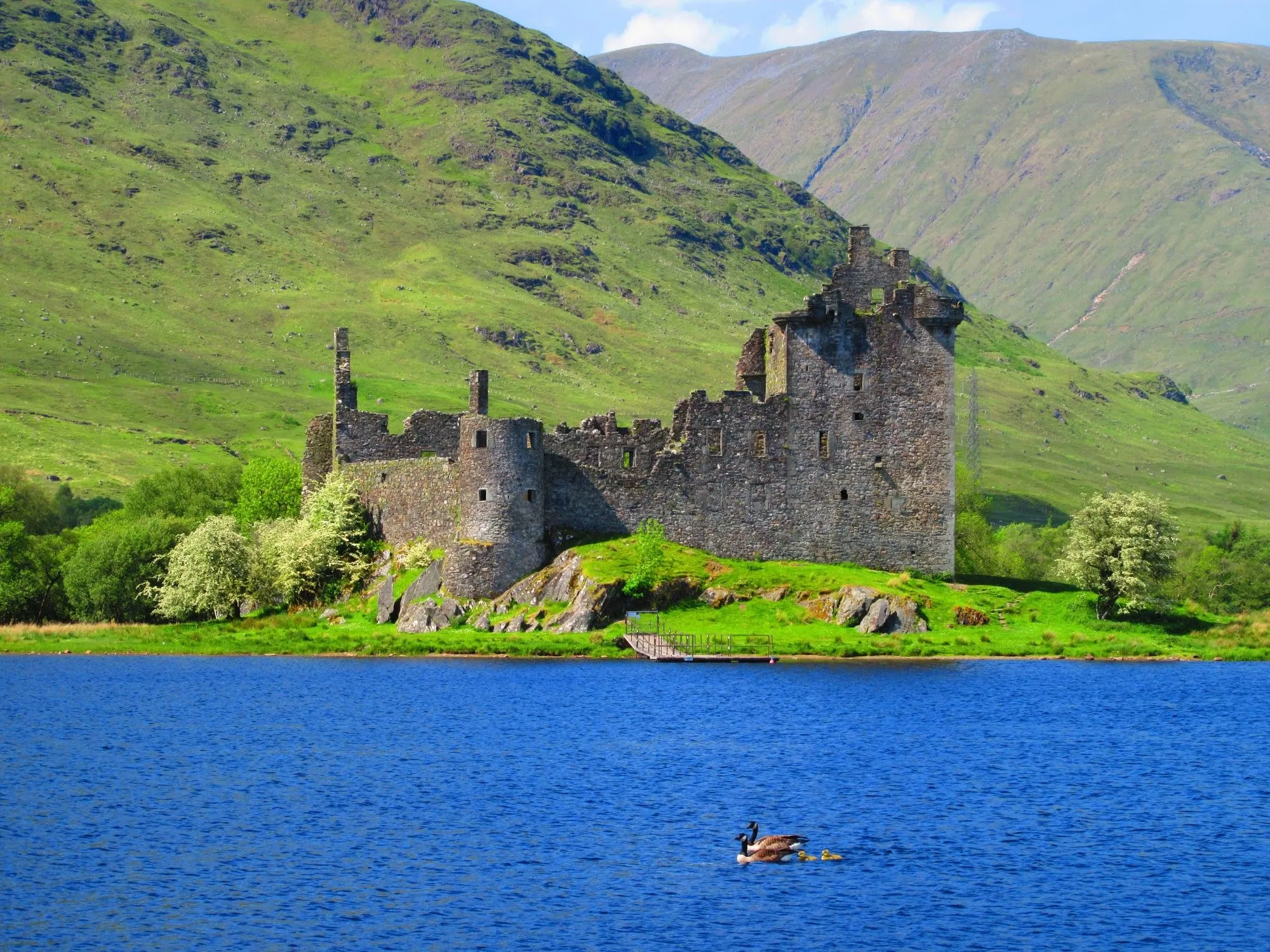 A beautiful and well-composed image of a historic castle ruin situated on the edge of a vibrant blue lake, backed by lush green mountains under a bright sky.