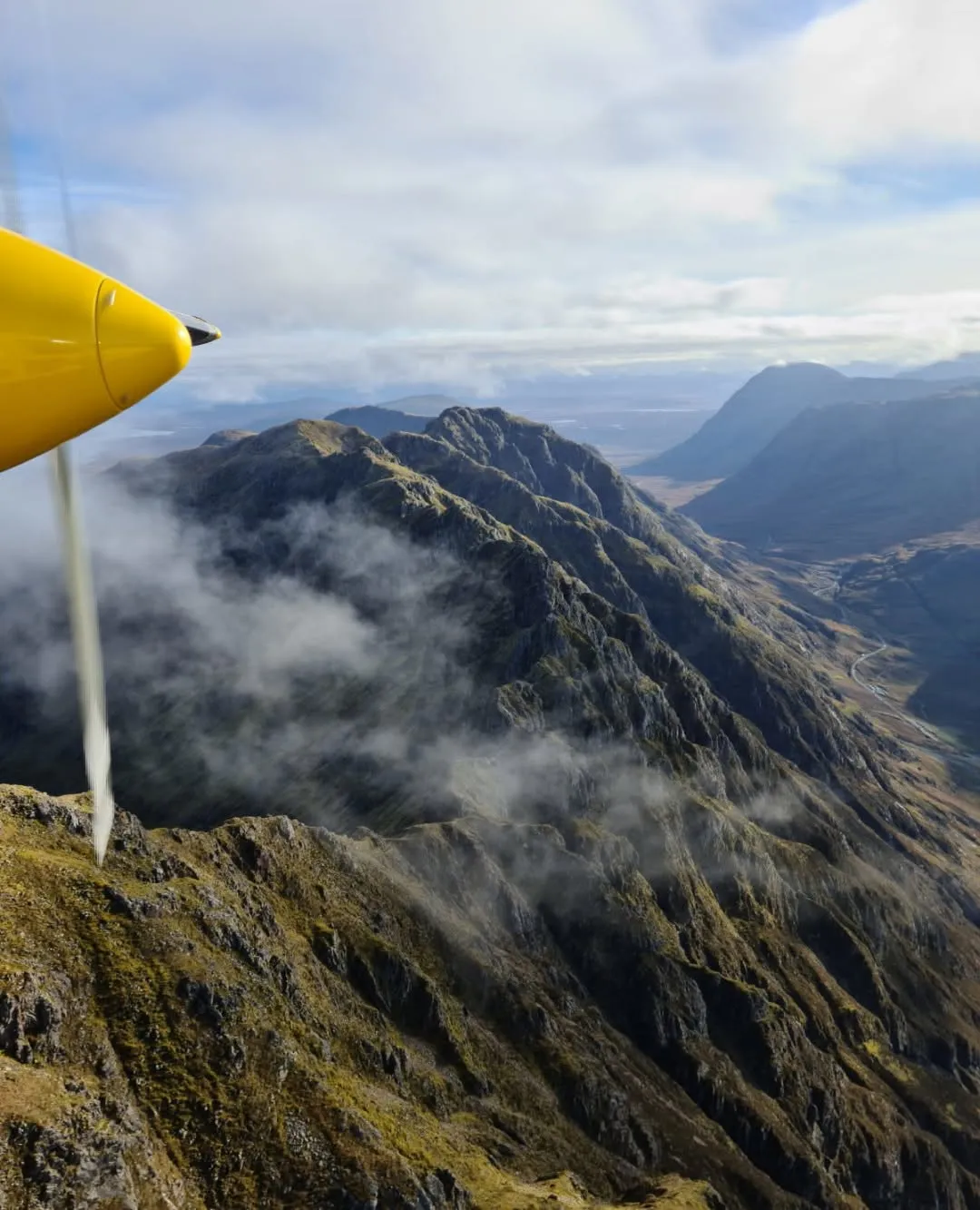 An aerial view showcasing rugged mountain ranges partially shrouded in clouds, with a bright yellow propeller of an aircraft in the foreground.