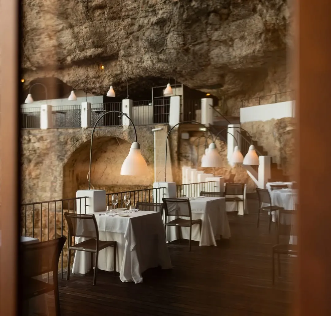 A captivating view of a restaurant interior, uniquely built within a natural cave or rock formation. Multiple dining tables, draped with white tablecloths and paired with chairs, are artfully arranged and illuminated by modern pendant lights hanging from elegant arching stands. The rugged, textured rock walls and ceiling of the cave create a distinctive and atmospheric backdrop, complemented by railings and walkways that suggest different levels within the space.