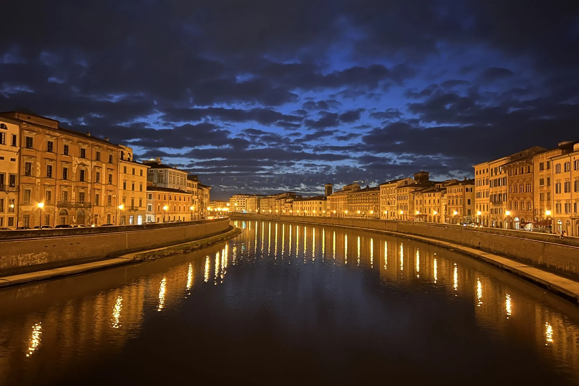 An editorial style image captured at night, showcasing a city river flanked by illuminated buildings on both sides, with their lights reflecting prominently on the water's surface under a dramatic cloud-filled sky.