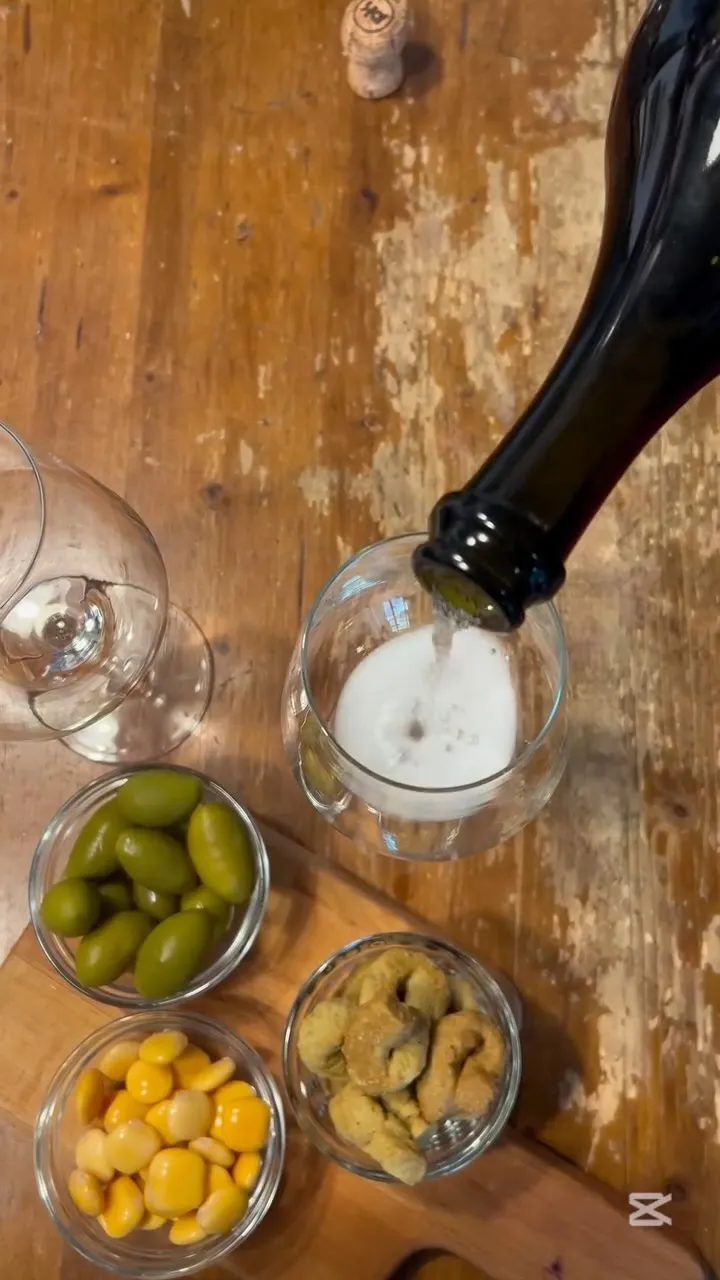 An overhead shot of sparkling wine being poured into a glass, with a bowl of olives and another empty glass on a wooden table.