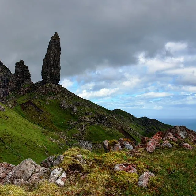 The image prominently features a majestic, tall rock formation set against a dramatic sky, surrounded by lush green hills. The foreground includes mossy rocks, adding texture and depth to the scene. In the distance, a glimpse of the ocean is visible.
