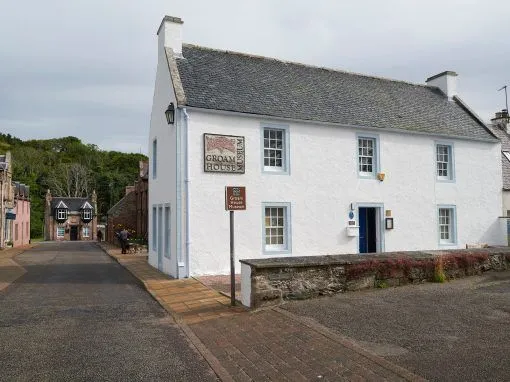 An exterior view of the Groam House Museum, a white historic building with blue window frames and a grey roof, situated on a street with a pedestrian path leading towards other distant buildings.