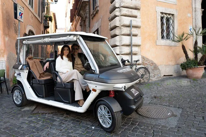 A man and a woman are seated in a white electric tour vehicle on a cobblestone street, surrounded by historic buildings.