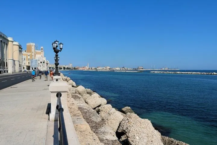 The image features a well-lit and vibrant coastal scene with a clear blue sky and deep blue sea. The foreground elements, including the lamppost and the promenade, are sharp, and the colors appear accurate and realistic. The composition is generally good, drawing the eye along the waterfront towards the distant city. The distant cityscape is less sharp, but this is typical for a wide scenic view and does not significantly detract from the overall quality. Key elements like the lamppost and the expansive sea are in focus.
