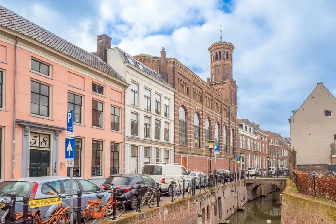 An inviting street view in a European city, showcasing a row of charming buildings, a tranquil canal, and a bustling street with parked cars and bicycles under a bright, cloudy sky. The architecture, including a prominent tower, is well-preserved, and the scene reflects a typical urban atmosphere.