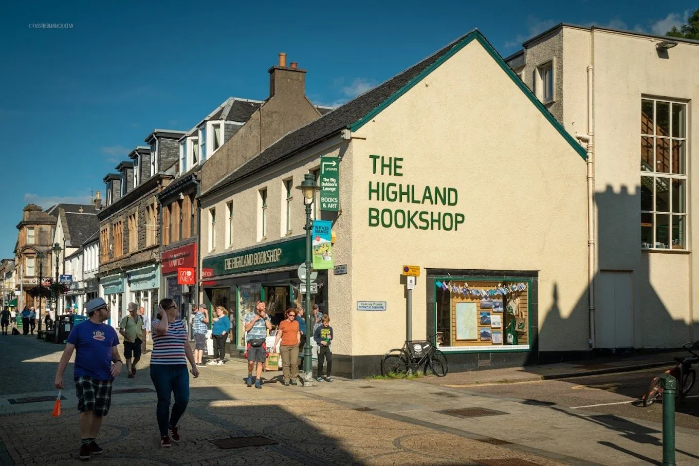 An exterior view of 'The Highland Bookshop' on a sunny day, with people walking on the cobblestone street in front of it. The building is a prominent feature, rendered in light yellow with green trim and text.