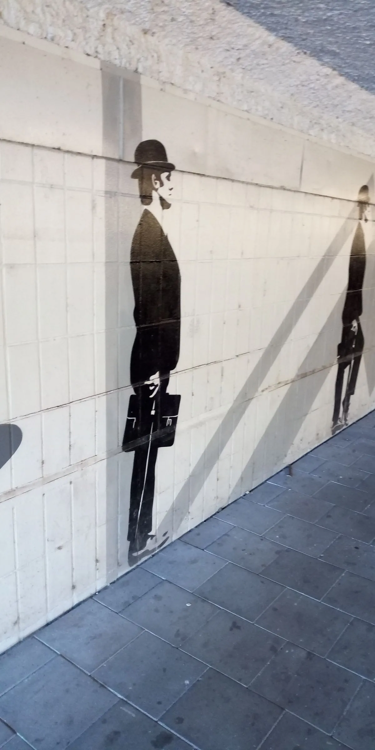 An urban scene featuring black stencil art of two figures, possibly businesspeople, holding briefcases, against a tiled wall. The scene is characterized by strong diagonal shadows crossing the surface.