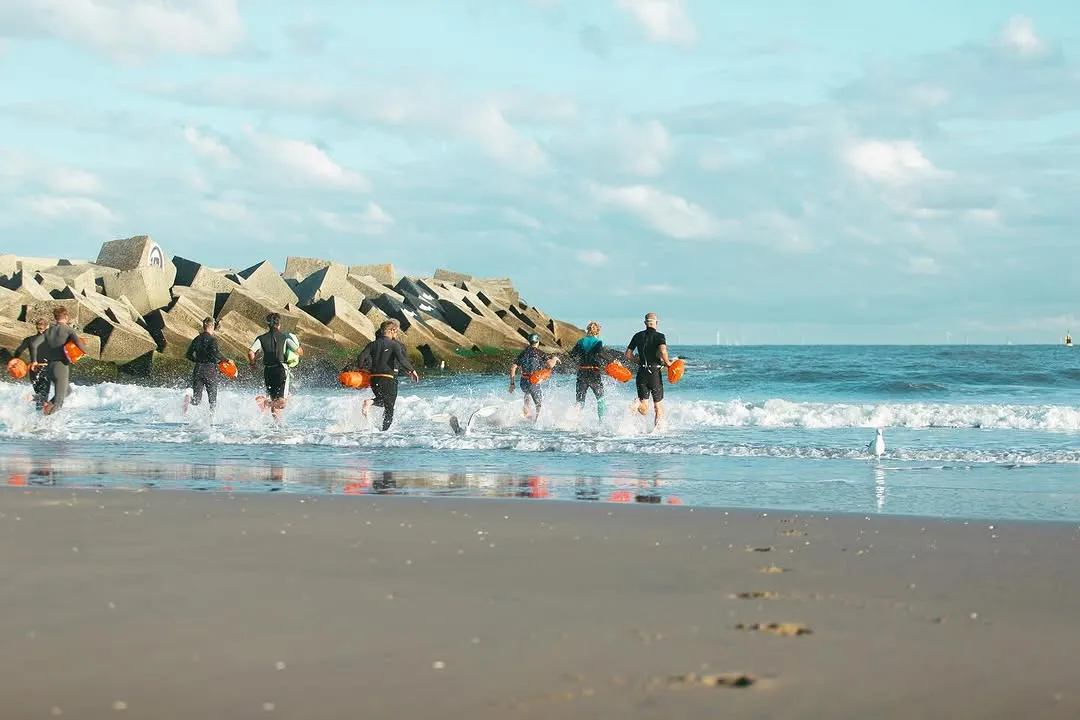 A group of people wearing wetsuits and carrying orange buoys run into the sea from a sandy beach, with a concrete breakwater in the background under a partly cloudy sky.