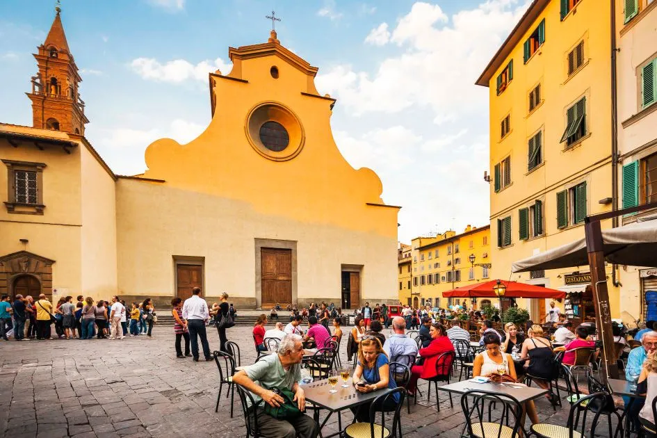 An outdoor scene depicting a lively city square with a prominent historic church building in the background. People are gathered, some walking, others seated at outdoor cafes, enjoying the atmosphere under a clear sky. The architecture is characterized by warm-toned buildings.