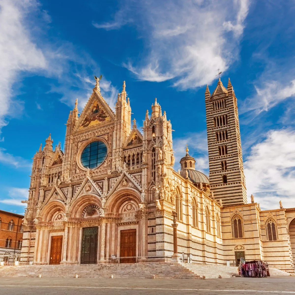 A grand exterior view of the Siena Cathedral (Duomo di Siena) under a bright blue sky with wispy clouds. The intricate Gothic facade with its detailed marble patterns and statues is prominently displayed, showing the main entrance and the striped bell tower.