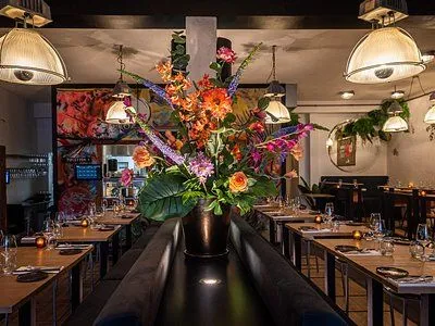 An editorial-style image capturing a restaurant interior with set tables, ambient lighting, and a prominent, vibrant floral centerpiece. The scene conveys a warm and inviting atmosphere.