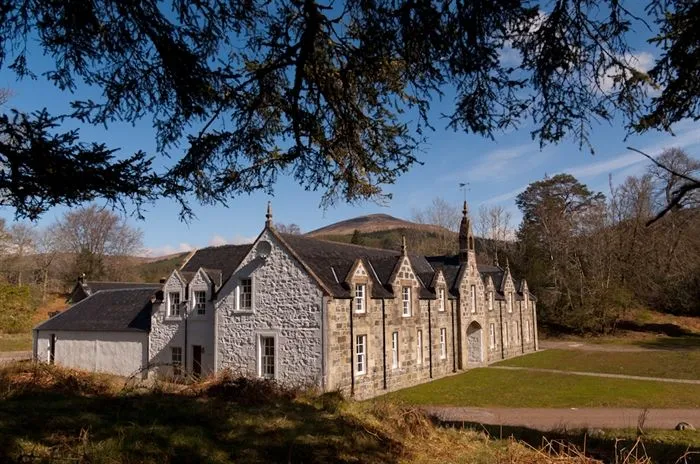 A well-composed photograph of a traditional stone building set within a natural landscape. The building is well-lit by natural light, showcasing its architectural details against a clear blue sky and distant hills.