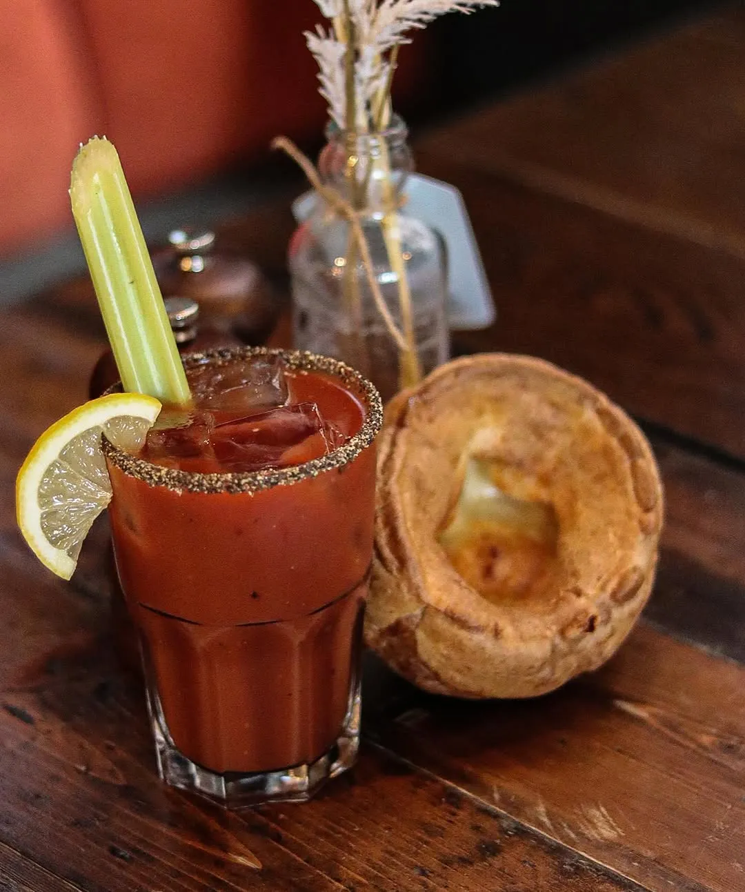A Bloody Mary cocktail garnished with celery and a lemon slice, served alongside a Yorkshire pudding on a wooden table.