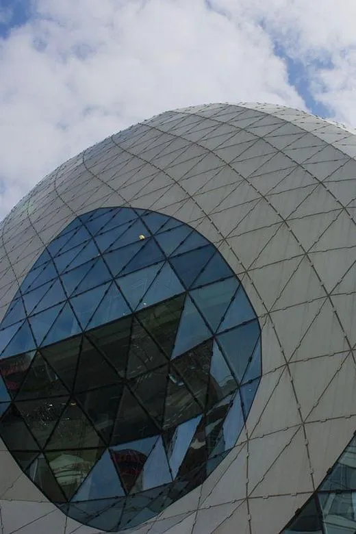 The image showcases a modern architectural structure with a curved, geodesic design, composed of intricate triangular glass and solid panels. It is very sharp, well-lit with natural light, and photo-realistic, accurately rendering the colors and textures of the building material. The composition is focused on the architectural detail, presenting the unique pattern and form clearly.