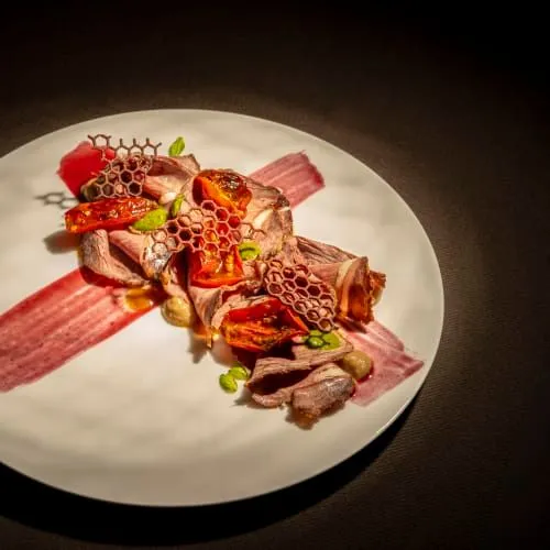 This is a professionally taken close-up image of a gourmet meat dish. The composition is excellent, centering the beautifully arranged food on a clean white plate. The lighting is dramatic and effective, expertly highlighting the textures and vibrant colors of the dish, especially the meat, sun-dried tomatoes, and unique honeycomb-like crisps. All key elements of the food are in sharp focus, contributing to a high degree of photo-realism and visual appeal. The colors appear accurate and appealing. The dark background ensures the food stands out as the sole subject, creating a sophisticated and appetizing presentation.