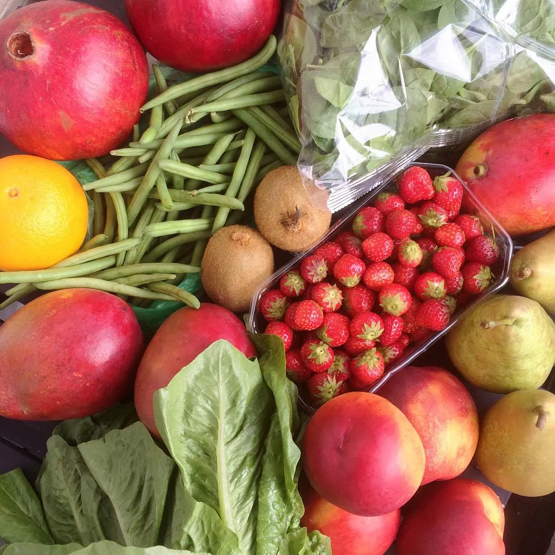 An overhead shot displaying a vibrant assortment of fresh fruits and vegetables, including pomegranates, green beans, kiwi, strawberries, mangoes, lettuce, peaches, and pears. The composition is full and highlights the natural colors and textures of the produce.