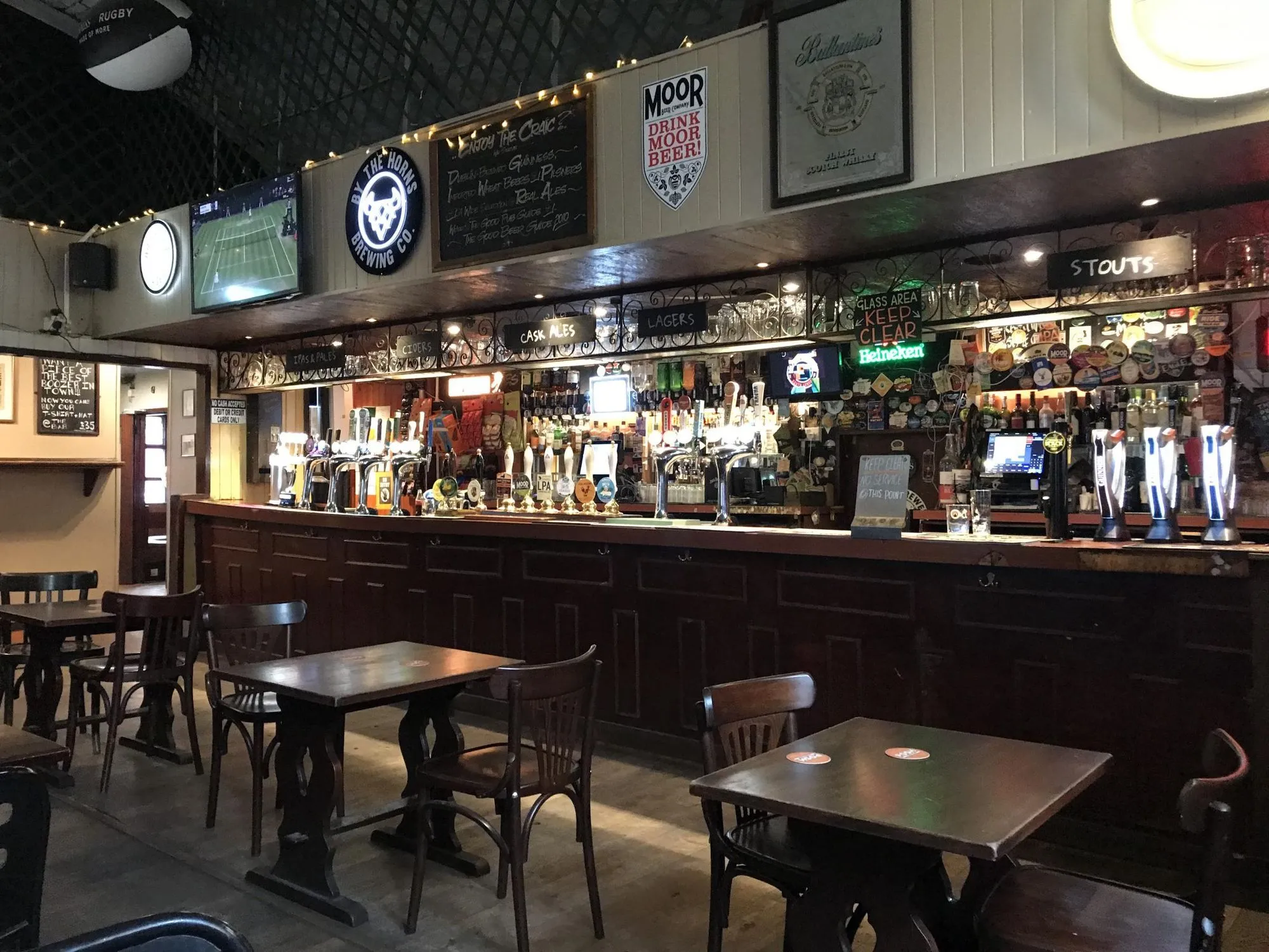 The image captures a well-composed interior of a bar, showcasing the full bar counter with taps, various signs, and seating in the foreground. The lighting is appropriate for a pub atmosphere, contributing to a natural look.