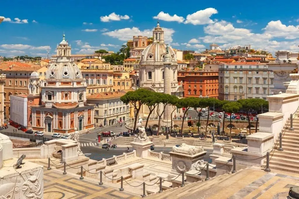 An editorial-style image capturing a vibrant cityscape under a bright, clear sky. The scene features grand historical buildings with prominent domes, intertwined with lush green trees. In the foreground, ornate stone steps and railings lead the eye towards a bustling street with some vehicles and pedestrians. The overall impression is one of architectural beauty and a lively urban environment, characteristic of a European historical city.