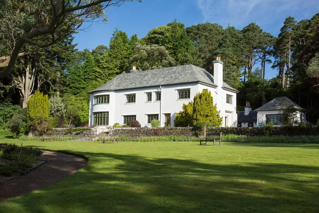 An exterior view of a large, white residential house with a grey roof, surrounded by a lush green lawn and dense trees under a clear blue sky.