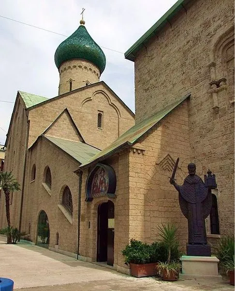 An exterior view of a light-colored brick church or cathedral with a prominent green dome, featuring a statue in the foreground near the entrance. The image captures architectural details and some surrounding foliage.