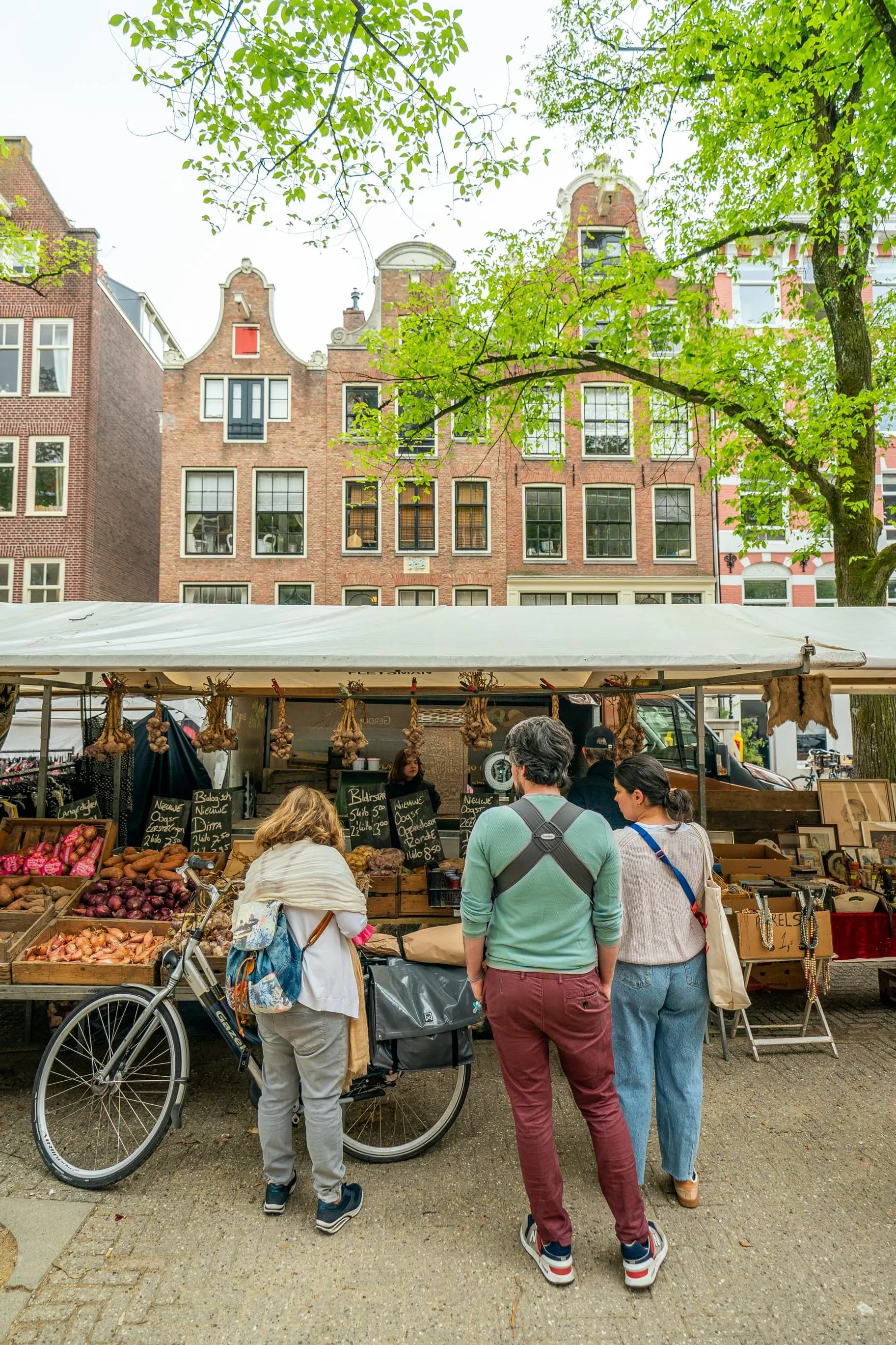 An outdoor market scene with a stall selling produce and several people browsing. Historic Dutch buildings are visible in the background, with a tree providing a touch of greenery.
