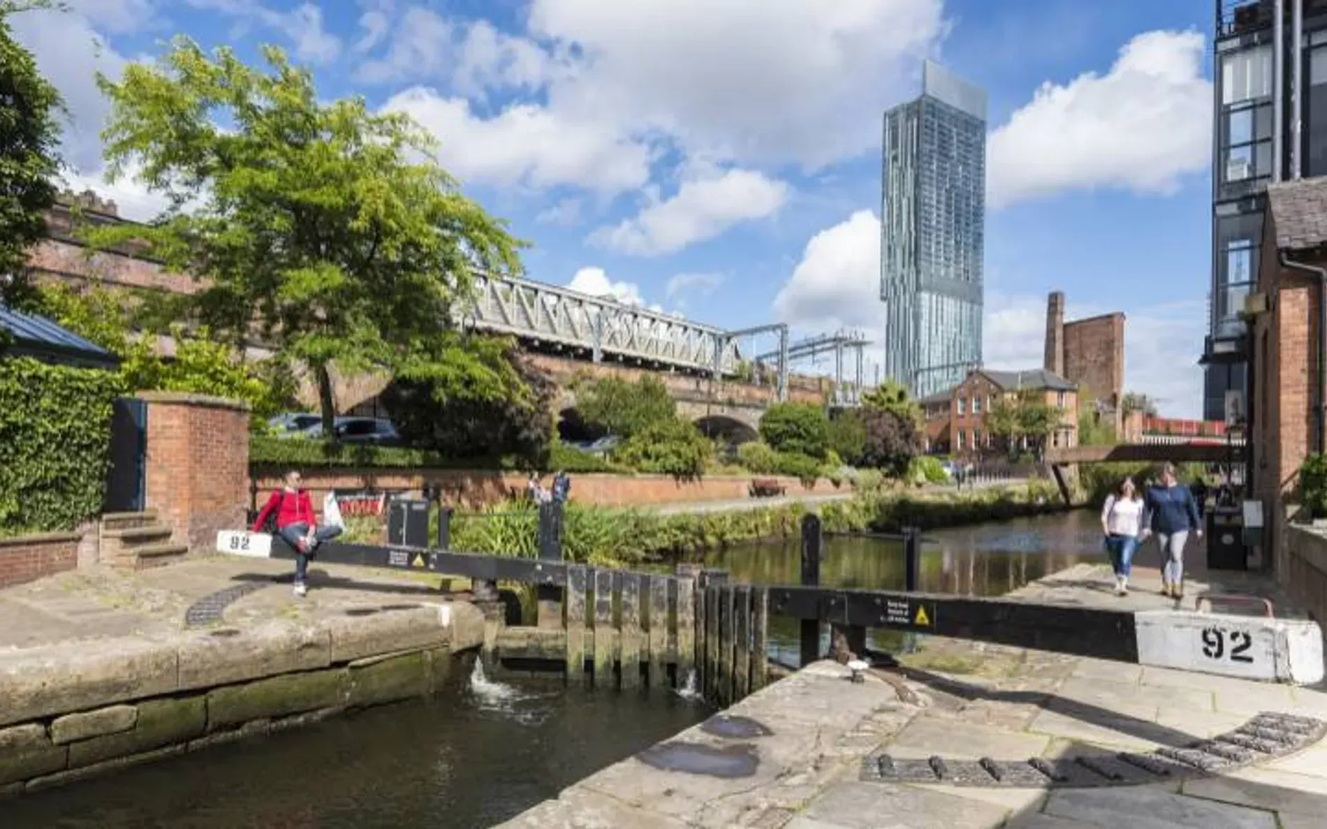 An editorial-style image capturing an urban canal scene under clear skies, featuring a modern skyscraper alongside older architecture and a bridge, with a canal lock in the foreground.