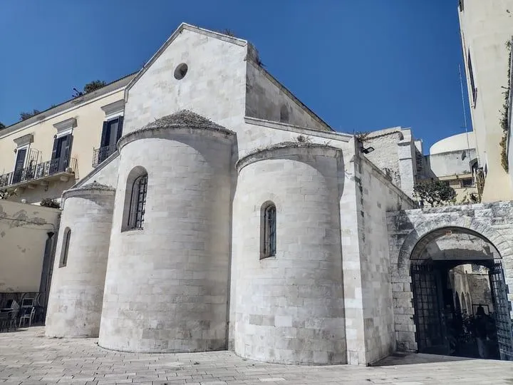 An exterior view of a historic white stone building, likely a church or an ancient structure, bathed in bright sunlight under a clear blue sky. The image focuses on the intricate architectural details, including rounded sections and narrow windows.