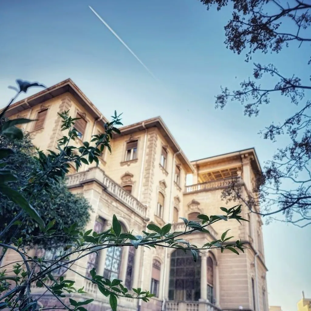 An ornate building with classic architecture, viewed from a low angle, partially framed by lush green foliage. The sky above is clear blue with a faint contrail.