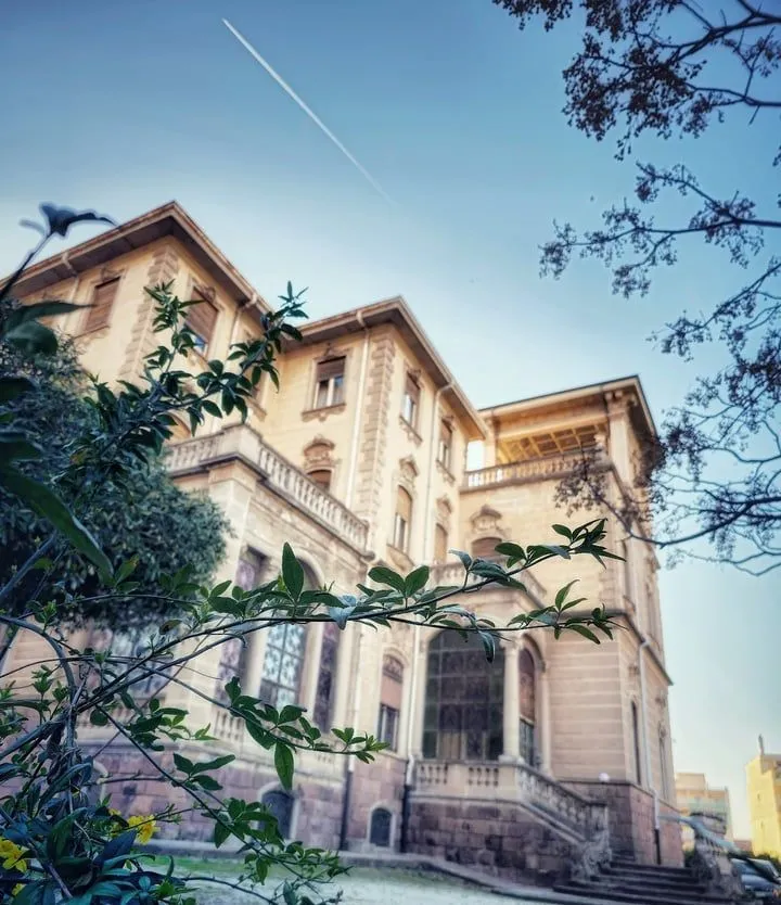 An ornate building with classic architecture, viewed from a low angle, partially framed by lush green foliage. The sky above is clear blue with a faint contrail.