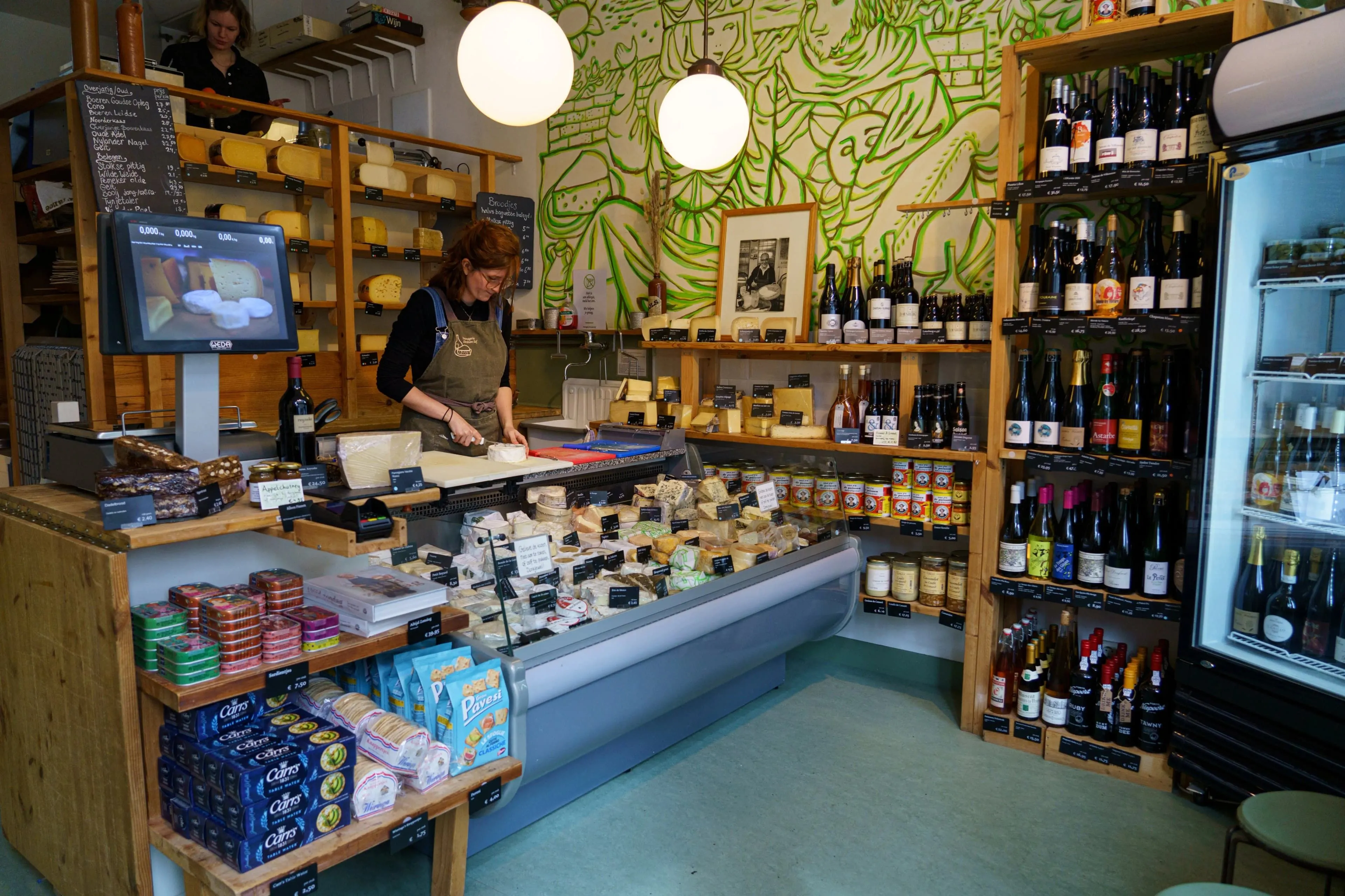 A well-lit, sharp, and photo-realistic editorial image depicting the interior of a specialty cheese and wine shop. A staff member is seen preparing cheese behind a counter filled with various cheese selections, while shelves stocked with bottles and other gourmet products line the walls. A vibrant green mural decorates the back wall.