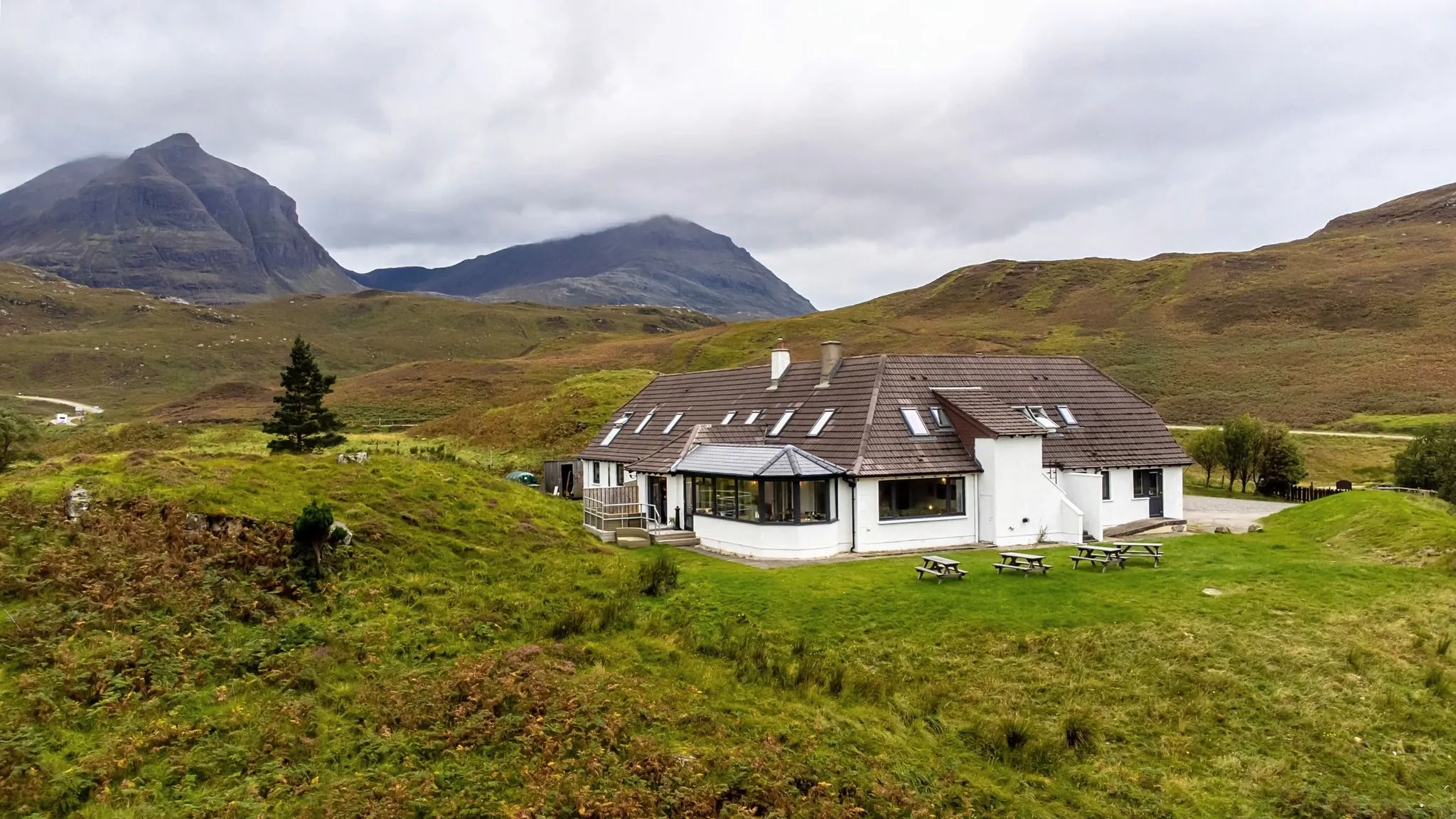 A white building with a brown roof stands prominently in a vast green and brown hilly landscape under an overcast sky, with large mountains visible in the background. Several wooden picnic tables are on the grassy area in front of the building.