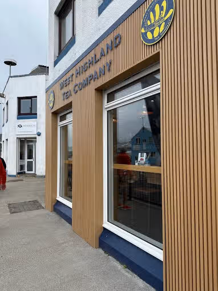 The image shows the exterior of the 'West Highland Tea Company'. The composition is decent, but the image isn't perfectly sharp. The lighting appears natural, possibly overcast, which results in even illumination but lacks strong highlights or shadows. Color accuracy seems reasonable. It's fairly photo-realistic. The key elements, such as the company name and the building's facade, are generally in focus. However, the overall image quality could be improved with better sharpness and more dynamic lighting.