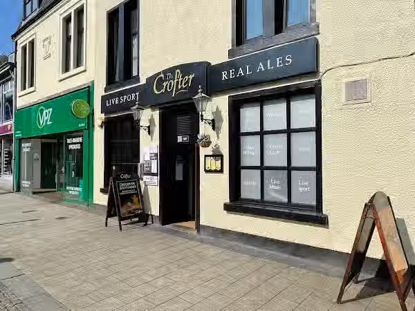 The image shows the exterior of a pub called 'The Crofter'. The composition is decent, capturing the building's facade. The lighting is acceptable, although slightly harsh, leading to some blown-out highlights. The sharpness is reasonable, but not exceptional. The colors appear accurate. The image is photo-realistic. The key elements, such as the pub's name and signage, are in focus. The image is well-composed and doesn't contain any unwanted elements. The image is an advertisement for the pub.