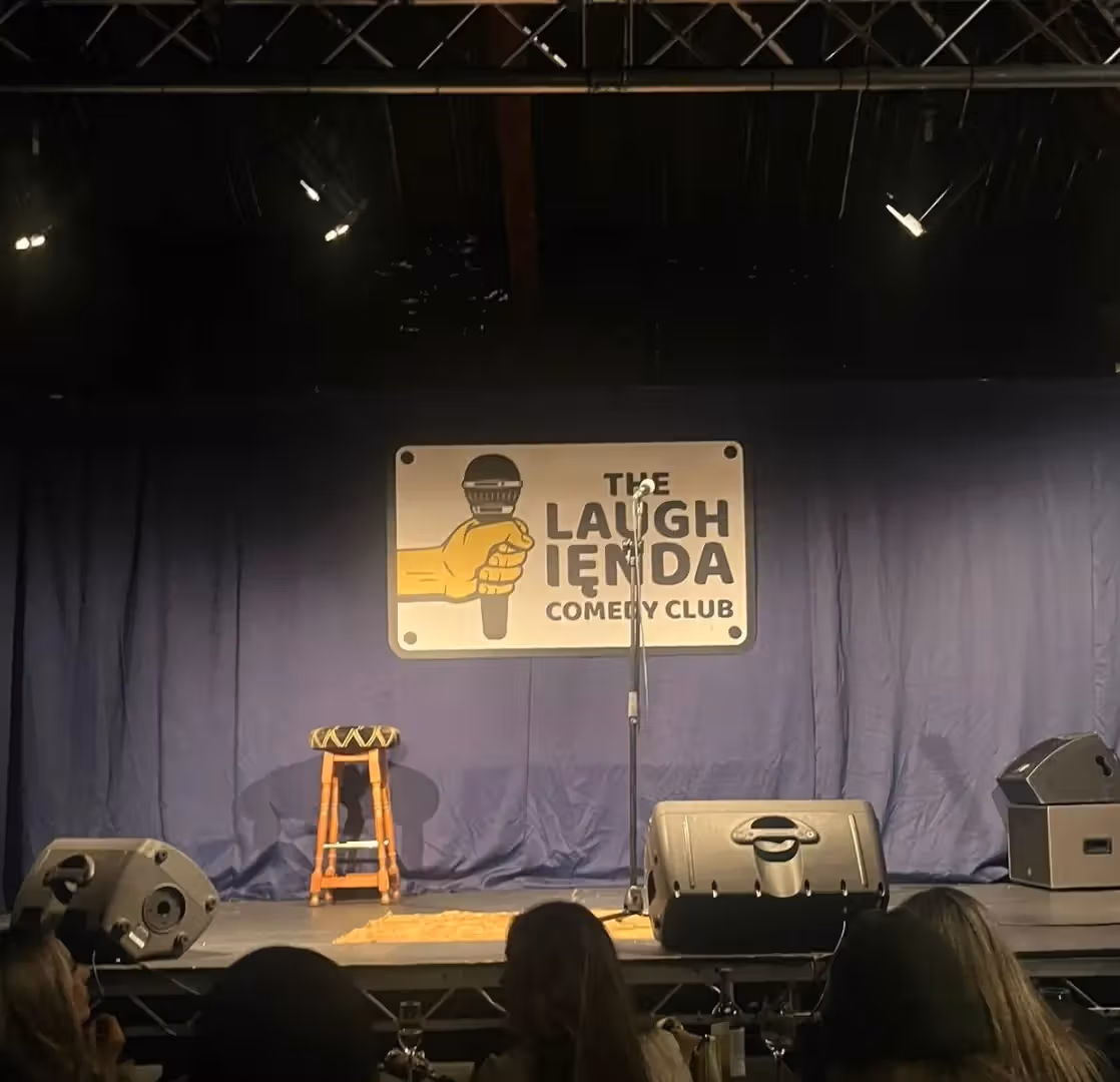 The image shows a stage at 'The Laughienda Comedy Club'. The composition is somewhat cluttered with speakers and the backs of peoples heads in the foreground. The lighting is dim and uneven. Sharpness is acceptable but not great. Color accuracy seems reasonable. It's not particularly photo-realistic. The key elements (logo, microphone) are reasonably in focus, but the overall image quality is impacted by the poor lighting and composition.