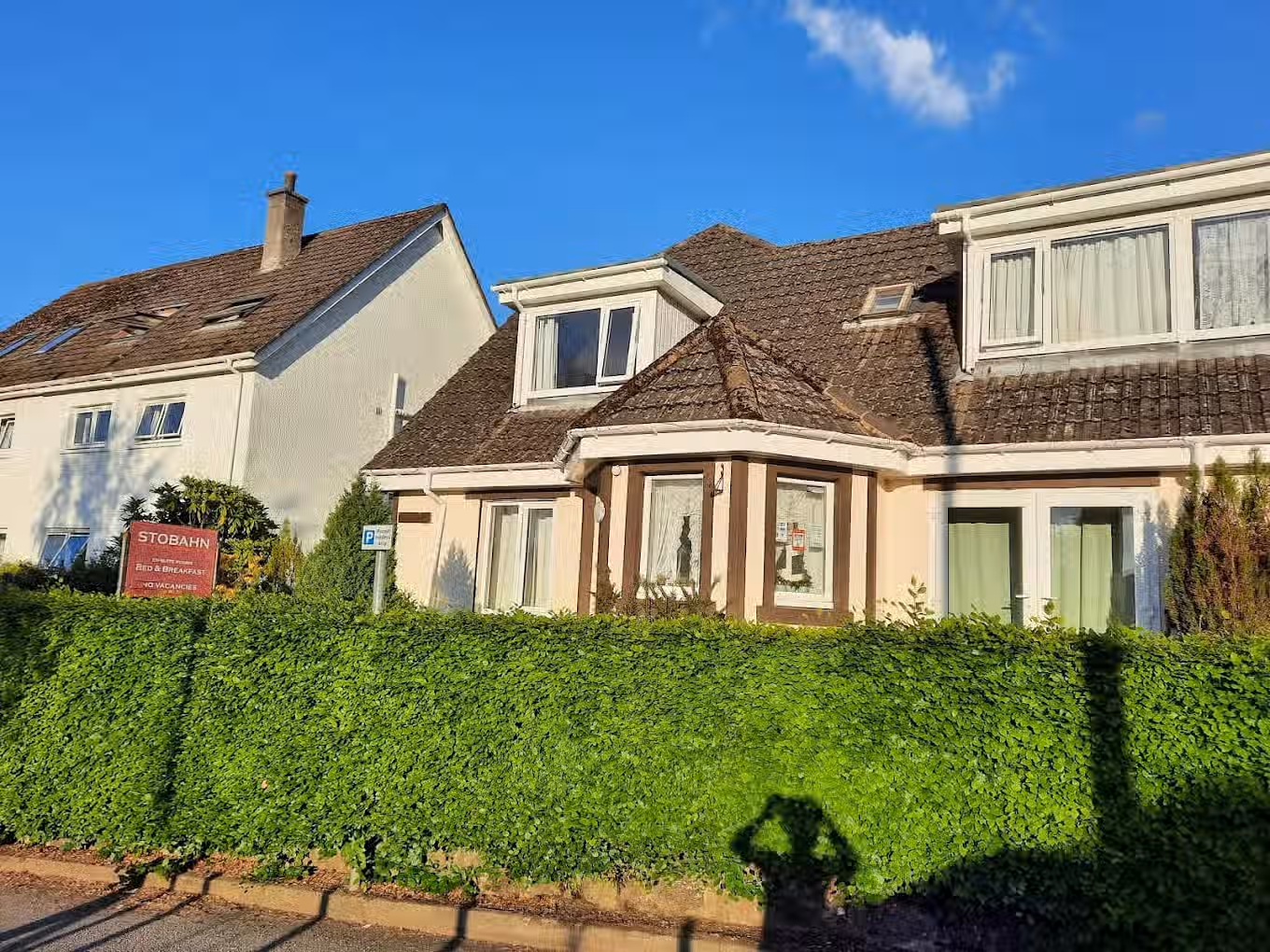 The image shows the exterior of a house. The composition is acceptable, but the shadow of the photographer at the bottom is an unwanted element. The lighting is good due to the daytime setting, and the colors appear accurate. However, the sharpness could be improved, and the photo-realism is average. The key elements, such as the house's facade, are mostly in focus. The shadow of the photographer impacts the overall quality.