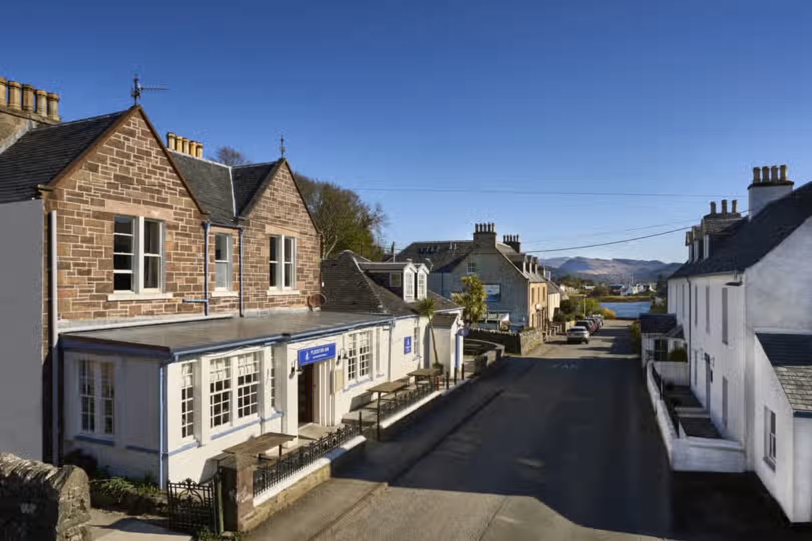 The image is well-composed and shows an exterior view of buildings and a street. The lighting is good, with a clear blue sky. The image is sharp and color-accurate. The key elements, such as the buildings, are in focus. The photo appears realistic and doesn't contain any unwanted elements according to the provided criteria. The composition is pleasing and showcases the architecture effectively. The only minor drawback is the slightly harsh shadows, but overall the image quality is high.