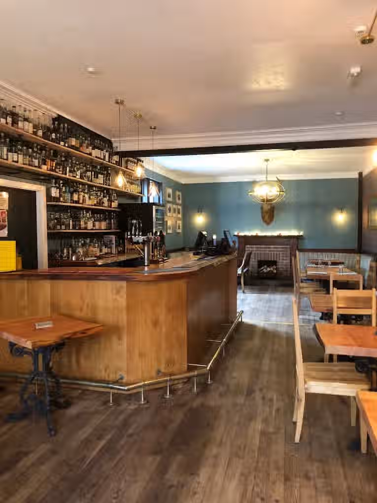 The image shows the interior of a bar. The composition is decent, capturing the bar area, tables, and a fireplace in the background. The lighting is adequate, though a bit flat. The sharpness could be improved. The colors appear accurate. The image is reasonably photo-realistic. Key elements like the bar and liquor bottles are generally in focus. The image is not cluttered and does not contain any unwanted elements. The composition is good, and the lighting is acceptable, but the sharpness and overall image quality could be better. The image is well-composed and lit, but the sharpness could be improved. The focus is on the bar area, but the background is also visible. The colors are accurate, and the image is photo-realistic.