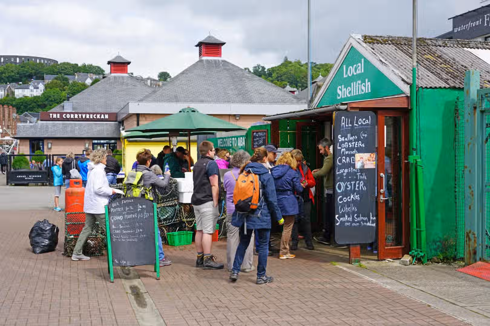 The image depicts a local shellfish market with people queuing. The composition is acceptable, but the scene is somewhat cluttered. The lighting is adequate, though a bit flat due to overcast conditions. Sharpness is moderate; details are visible but not exceptionally crisp. Color accuracy appears reasonable. The photo-realism is acceptable. Key elements like the menu boards and the storefront are in focus. The image is not a professionally taken close-up food image, but it is not visually unappealing or taken by a guest. It contains text (menu boards), which is allowed. The image is slightly cluttered, but this is somewhat expected in a market scene. Well-composed: 20/25, Well-lit: 12/15, Sharp: 15/20, Color-accurate: 8/10, Photo-realistic: 10/20, Key elements in focus: 10/10