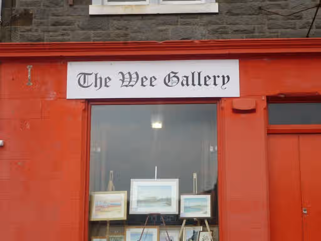 The image shows the exterior of 'The Wee Gallery'. The composition is adequate, but the lighting is somewhat flat and the sharpness could be improved. The color accuracy appears reasonable, but the overall image lacks a sense of photo-realism. The key elements, such as the gallery sign and the paintings displayed in the window, are reasonably in focus. The image is not particularly well-composed, and the overall aesthetic is somewhat bland. The image is not a professionally taken close-up food, selfie, promotional graphic, advertisement, poster, or text-based graphic, so it is not penalized for not being any of those. The image is not of half-eaten food, drinks that aren't full, or a cluttered environment, so it is not penalized for any of those. The image is not a visually unappealing close-up food image taken by guests, so it is not penalized for that.
