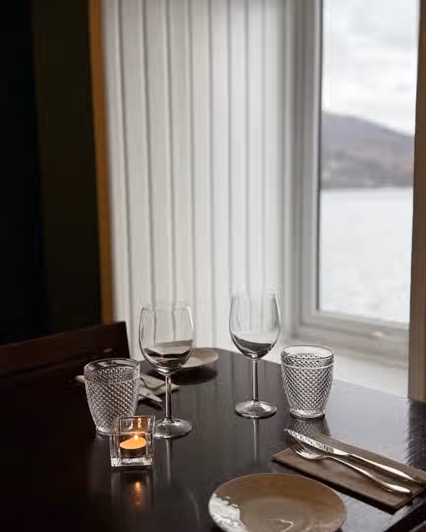 The image shows a well-composed table setting with wine glasses, water glasses, and a candle. The window view in the background adds depth. The lighting is soft and even, contributing to a pleasant atmosphere. The sharpness is adequate, and the colors appear accurate. The key elements, such as the glasses and candle, are in focus. It is not photo-realistic but it is well-shot. The image does not contain any unwanted elements according to the prompt's criteria.