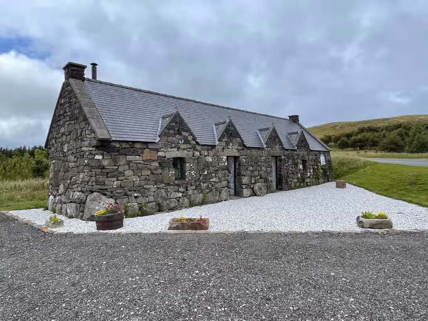 The image shows a stone building in a rural setting. The composition is decent, but could be improved. The lighting is adequate, though somewhat flat due to the overcast sky. Sharpness is acceptable, but not exceptional. Color accuracy appears good. The image is reasonably photo-realistic. Key elements, such as the building's facade, are in focus. Well-composed (20/25), Well-lit (12/15), Sharp (15/20), Color-accurate (8/10), Photo-realistic (15/20), Key elements in focus (5/10).