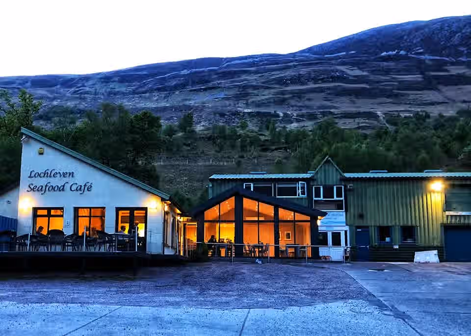 The image shows the exterior of a cafe with mountains in the background. The composition is decent, but the lighting is not ideal, as it appears to be taken during dusk or dawn, resulting in a somewhat dark image. The sharpness is acceptable, but not exceptional. Color accuracy seems reasonable, though the overall image has a slightly cool tone. The photo-realism is good, but the lack of optimal lighting detracts from it. The key elements, such as the cafe and the mountains, are generally in focus. The image is slightly underexposed. Well-composed / 25 = 18, Well-lit / 15 = 8, Sharp / 20 = 14, Color-accurate / 10 = 7, Photo-realistic / 20 = 12, Key elements in focus / 10 = 6