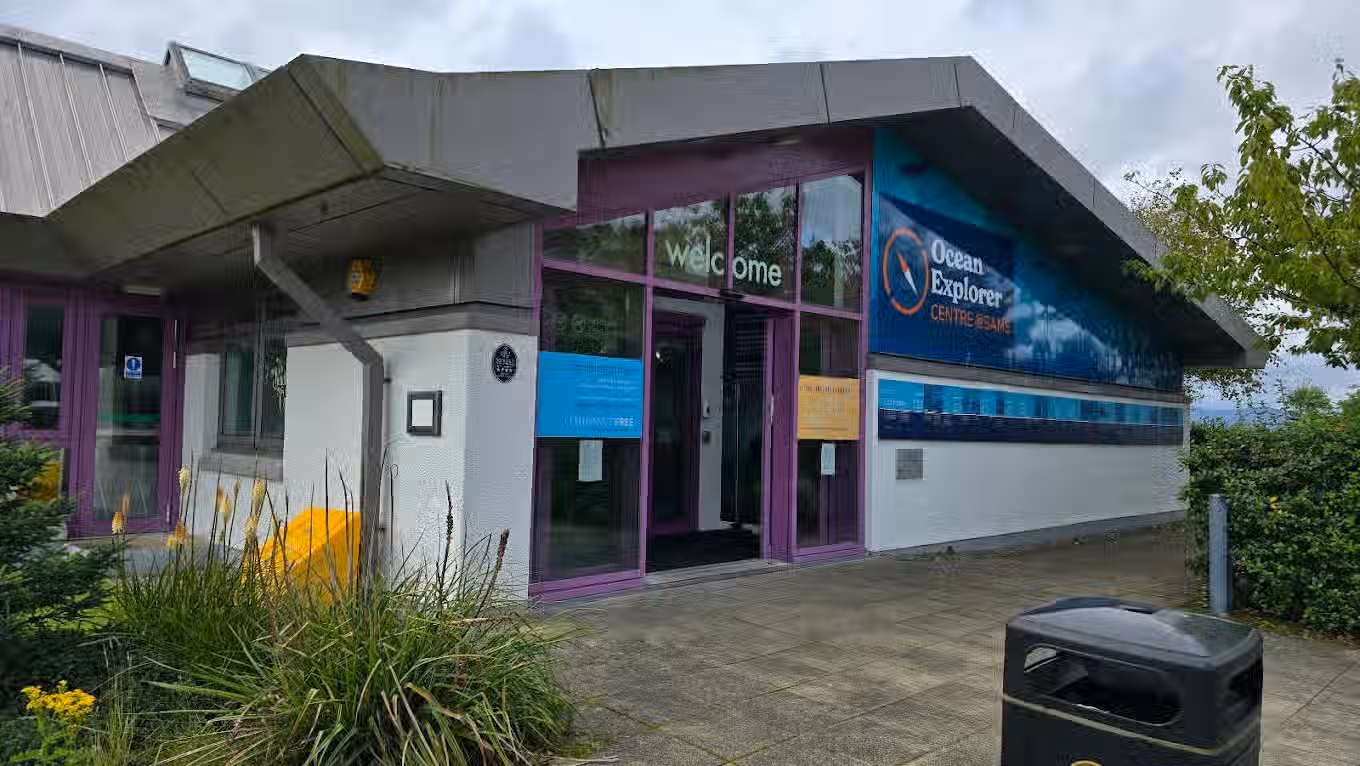 The image shows the exterior of a building, specifically the entrance to the Ocean Explorer Centre. The composition is adequate, but could be improved by focusing more on the entrance itself and less on the surrounding pavement and foliage. Lighting is somewhat flat, which impacts the overall visual appeal. Sharpness is acceptable but not exceptional, and the colors appear accurate. It's not particularly photo-realistic, but serves as a decent representation of the location. The key elements, such as the signage and entrance, are in focus. The image could be improved with better lighting and a more focused composition. The presence of the trash can on the right side of the image is a minor distraction. Based on the criteria, the image receives a score of 65. Strengths: - Clear depiction of the building entrance and signage - Accurate colors Weaknesses: - Flat lighting - Average sharpness - Could be better composed