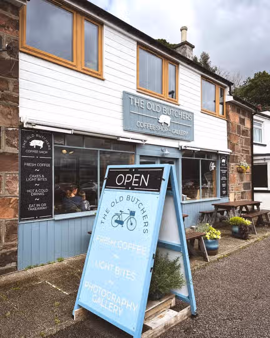The image shows the exterior of 'The Old Butchers' coffee shop and gallery. The composition is good, with the shopfront and 'Open' sign clearly visible. The lighting is adequate, though slightly overcast, which affects the overall brightness. The image is reasonably sharp, and the colors appear accurate. It has a photo-realistic quality. The key elements, such as the shop name and the 'Open' sign, are in focus. There are no unwanted elements present. The image serves as an advertisement for the business.