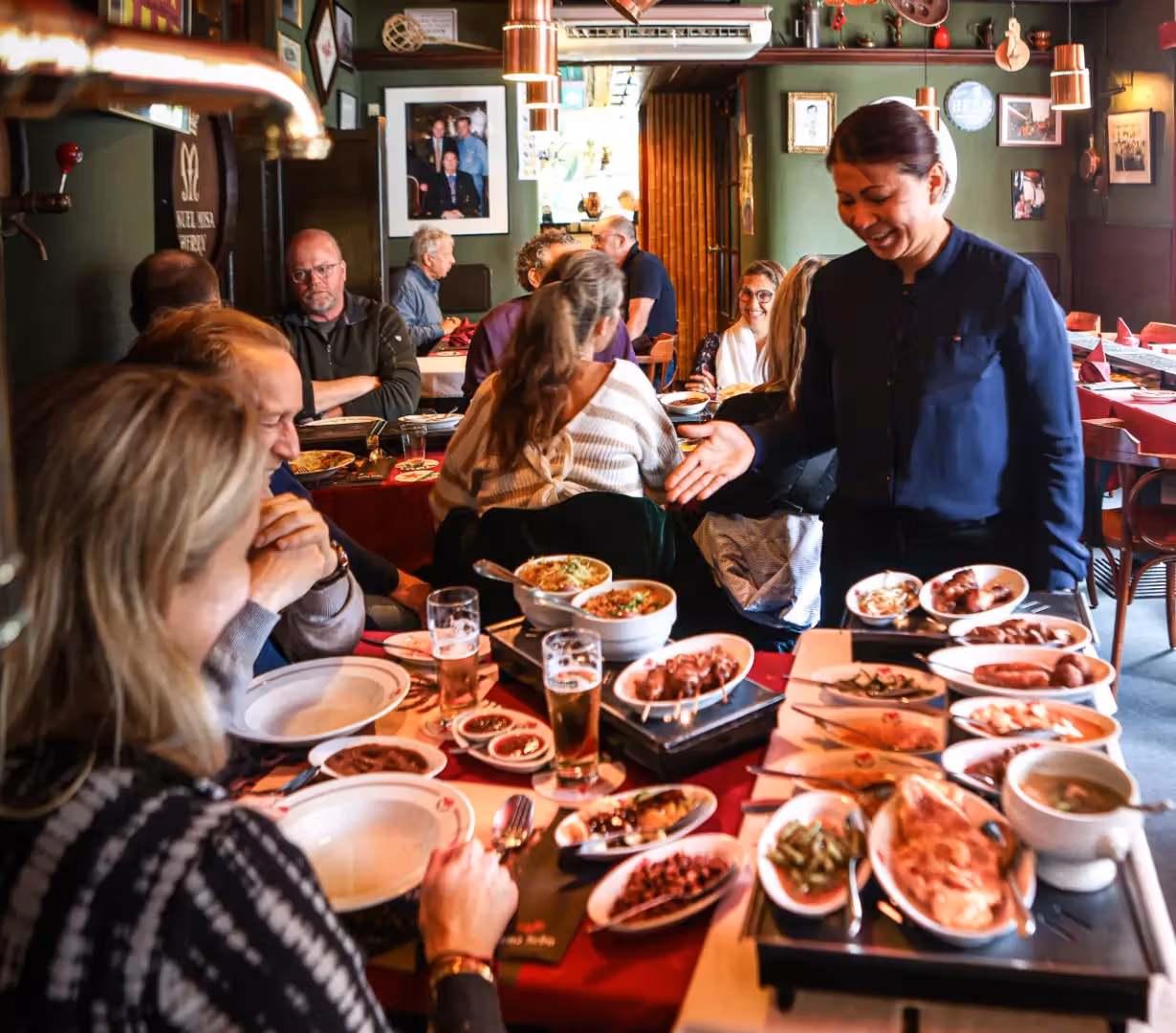 The image shows a restaurant scene with a waitress serving food to customers at a table. The composition is acceptable, but the lighting is somewhat uneven, and the sharpness could be improved. The colors appear relatively accurate. Some of the beer glasses aren't completely full. The key elements, such as the food and people, are mostly in focus, but the overall image quality is not exceptionally high. The image doesn't contain any watermarks or logos, and no half eaten food is visible. The environment is slightly cluttered but acceptable. The photo-realism is fair.