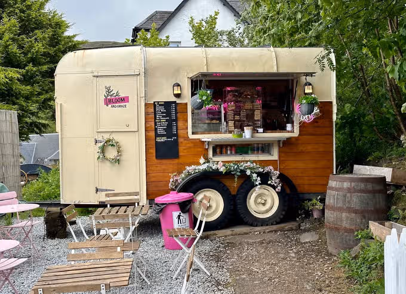 The image depicts a food truck called 'Bloom and Graze'. The composition is acceptable, but the presence of a pink trash can is visually distracting. The lighting is adequate, but the sharpness could be improved. The colors appear accurate. The image is reasonably photo-realistic. The key elements, such as the food truck itself and the menu board, are generally in focus. However, the overall impression is somewhat diminished by the trash can and the slightly cluttered environment.