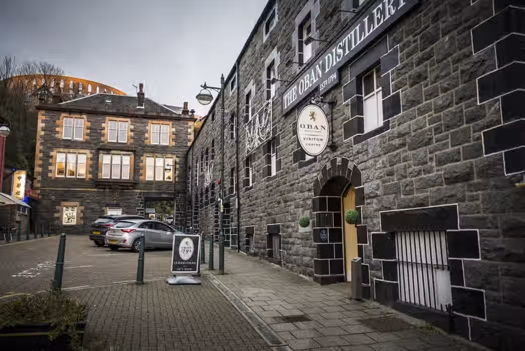 The image depicts the exterior of the Oban Distillery. The composition is decent, although the inclusion of parked cars detracts slightly. The lighting is somewhat flat due to the overcast conditions, impacting the overall vibrancy. Sharpness is acceptable, but not exceptional. Color accuracy appears reasonable. While not perfectly photo-realistic, it captures the scene adequately. Key elements like the distillery sign and entrance are in focus. The presence of cars is a minor detractor, but the image is otherwise acceptable. The overall image quality is good, but could be improved with better lighting and composition.