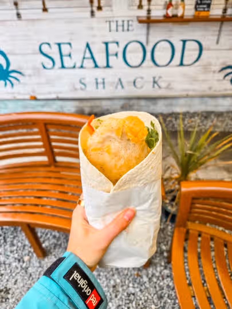 The image shows a seafood wrap being held in front of a 'Seafood Shack' sign. The composition is acceptable, but the lighting is somewhat flat. The image is reasonably sharp, and the colors appear mostly accurate. It doesn't appear overly photo-realistic, and the key element (the wrap) is in focus. However, the hand holding the wrap and the background are slightly distracting. The overall presentation is acceptable but not exceptional. It is not a professionally taken close-up food photo, but it doesn't fall into the 'unwanted elements' category. The image is acceptable, but could be improved with better lighting and composition.